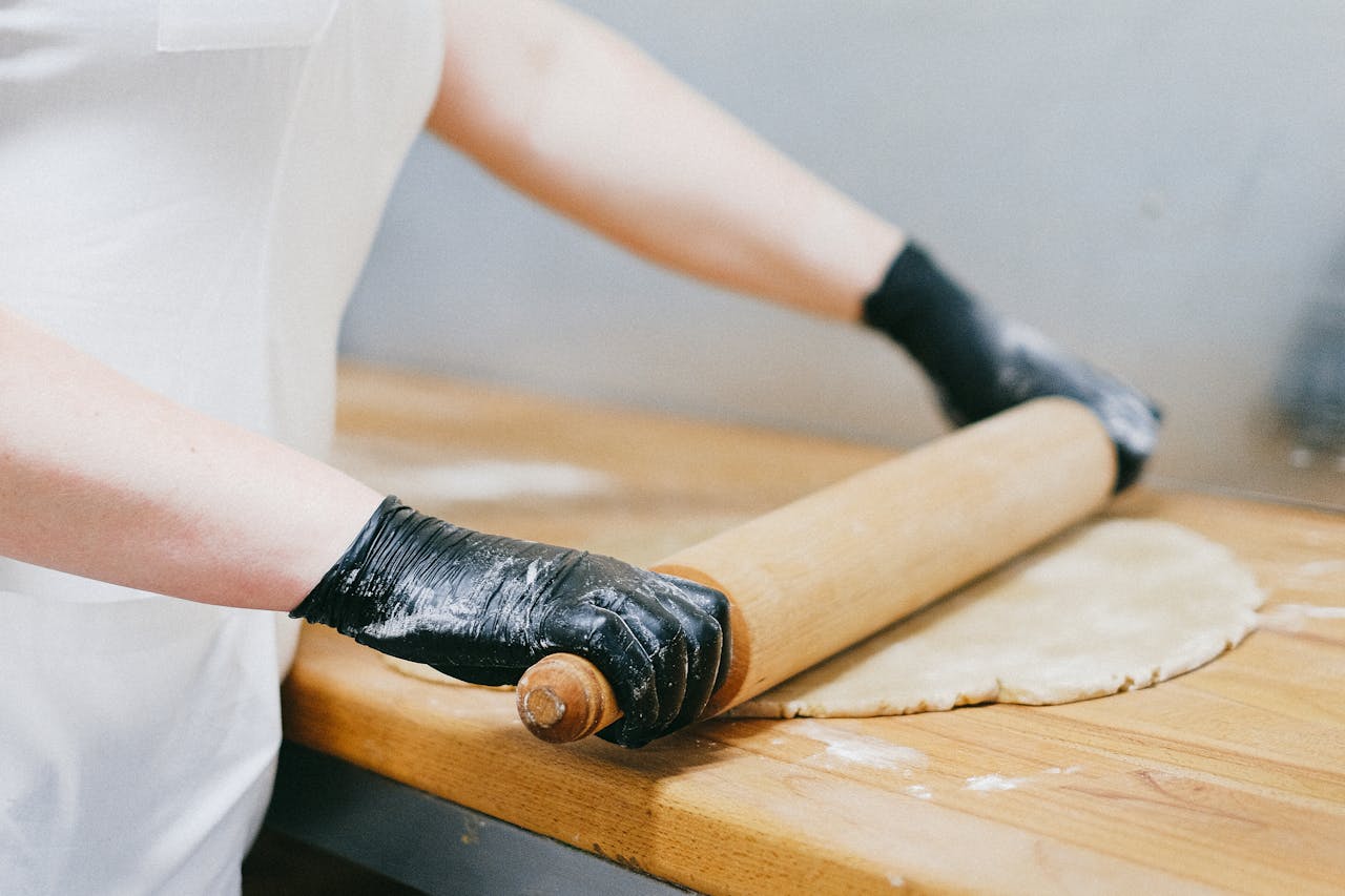 Close-up of a baker skillfully rolling dough with a wooden pin while wearing black gloves.